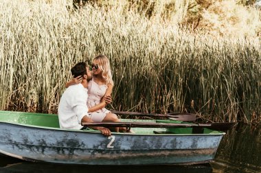 young couple hugging and kissing in boat on river near thicket of sedge clipart