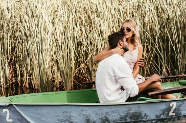 attractive young woman embracing boyfriend in boat on lake near thicket of sedge clipart