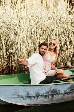 smiling man pointing with finger while sitting with girlfriend in boat near thicket of sedge clipart