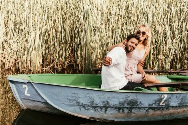 cheerful man pointing with finger while sitting with girlfriend in boat near thicket of sedge clipart