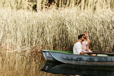 happy young couple embracing while sitting in boat on river near thicket of sedge clipart