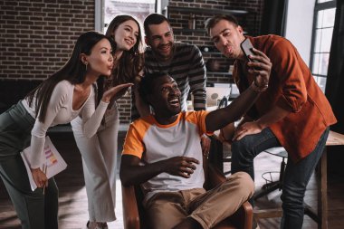 Mutlu Afro-Amerikan iş adamı akıllı telefonuyla selfie çekerken etrafı neşeli, çok kültürlü iş arkadaşlarıyla çevrili koltukta oturuyor.