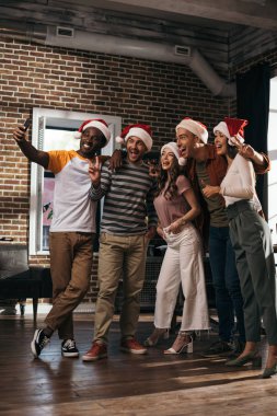 Mutlu Afro-Amerikan iş adamı, Noel Baba şapkalı neşeli çok kültürlü meslektaşlarıyla akıllı telefondan selfie çekiyor.