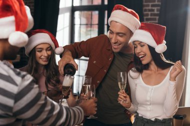 Neşeli iş adamı Noel Baba şapkalı mutlu meslektaşlarının kadehlerine şampanya dolduruyor.