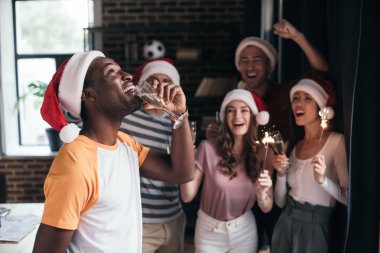 Neşeli Afro-Amerikan iş adamı Noel Baba şapkalı çok kültürlü meslektaşlarının yanında dururken şampanya içiyor.