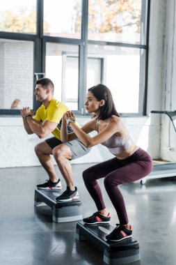Sporcu ve sporcu kadın spor merkezindeki platformlarda çömelme yapıyorlar.