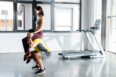 Spor merkezinde çömelen ve sporcu kadını omuzlarında tutan sporcunun yan görüntüsü