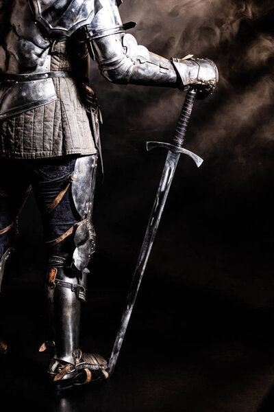 cropped view of knight in armor holding sword on black background 
