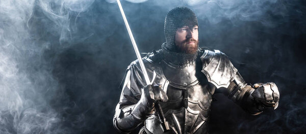 panoramic shot of handsome knight in armor looking away and holding sword on black background 