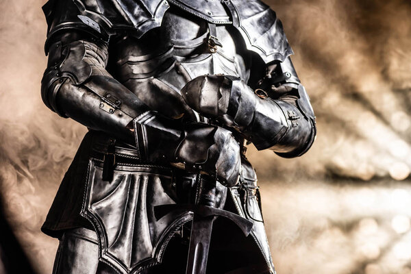 cropped view of knight in armor holding sword on black background