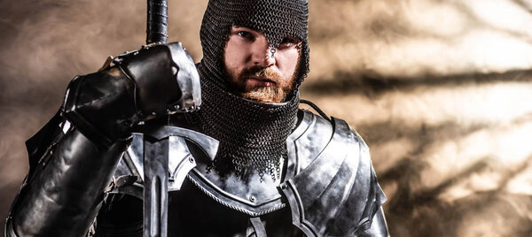 panoramic shot of handsome knight in armor holding sword on black background
