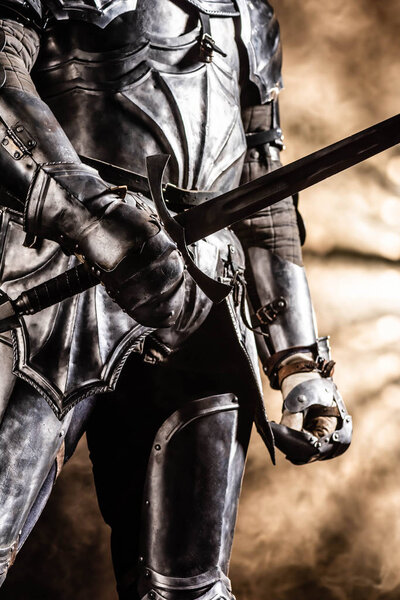 cropped view of knight in armor holding sword on black background