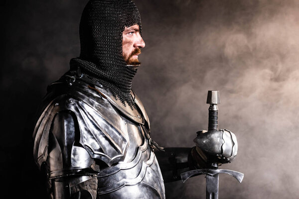 side view of handsome knight in armor holding sword on black background