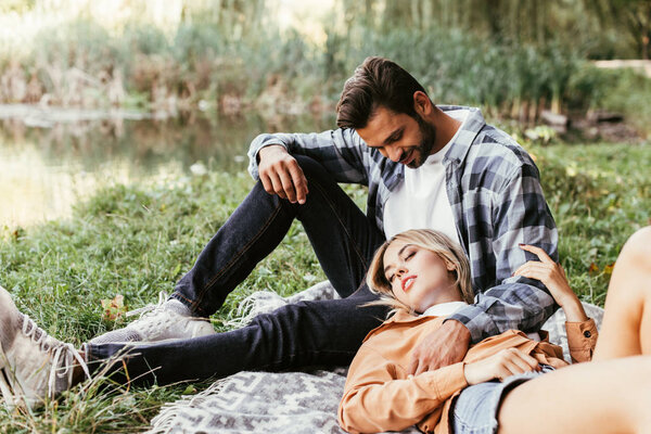 Красивый мужчина обнимает девушку, спящую на одеяле возле озера в парке
