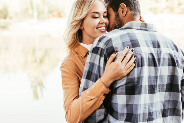 happy young woman embracing boyfriend while standing near lake in park