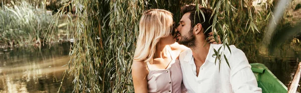 panoramic shot of happy young couple kissing while sitting in boat on lake