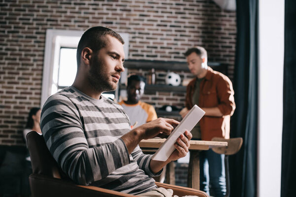 concentrated businessman using digital tablet while sitting in armchair near young multicultural colleagues