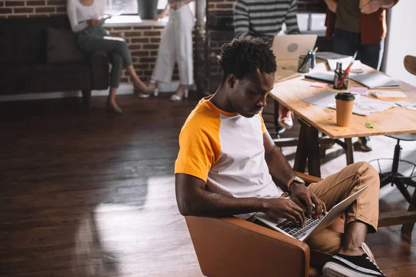 专心致志的非洲商人坐在写字台旁边的扶手椅上 一边用笔记本电脑一边喝咖啡一边走 — 图库照片