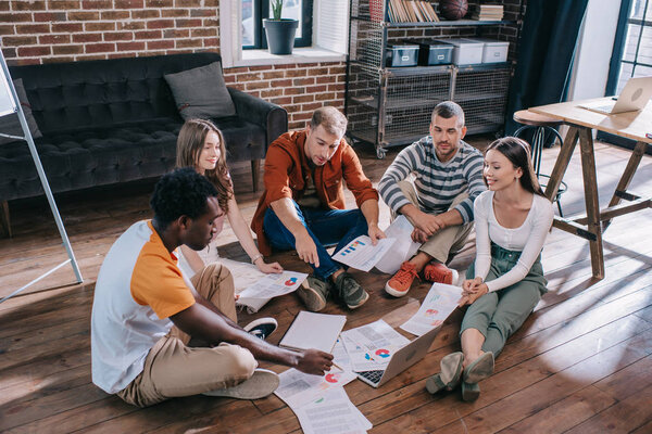 young businesspeople sitting on floor near documents with infographics and discussing business ideas