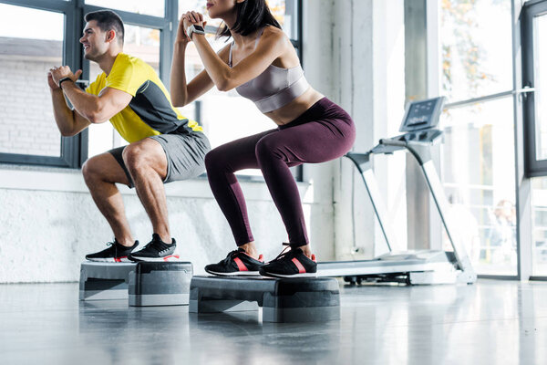 cropped view of sportsman and sportswoman doing squat on step platforms in sports center