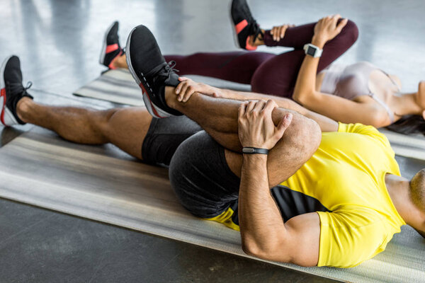 обрезанный вид спортсмена и спортсменки, растянувшейся на фитнес-ковриках в спорткомплексе
