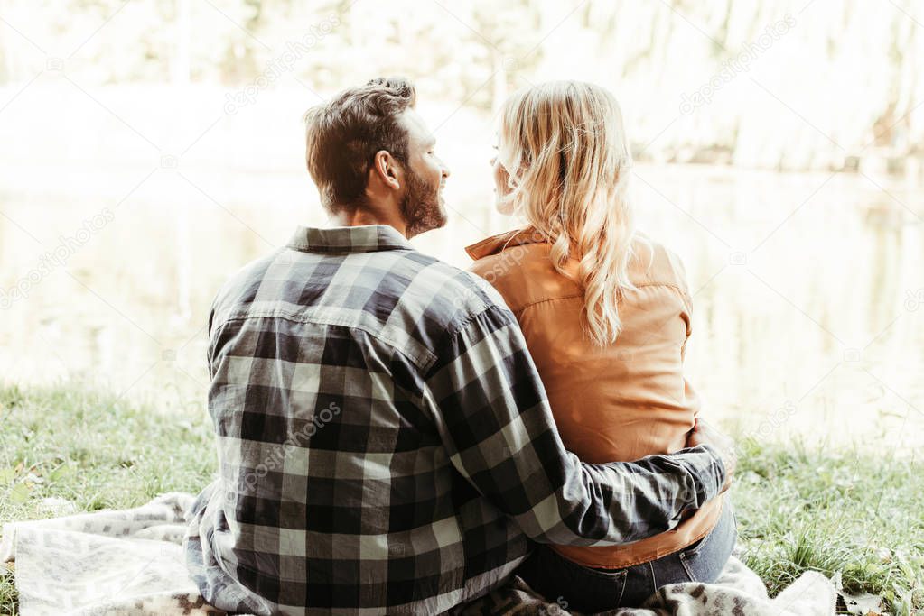 back view of young man embracing girlfriend while sitting near lake in park