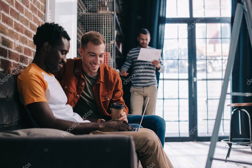 smiling african american businessman using laptop while sitting on sofa near cheerful colleague 