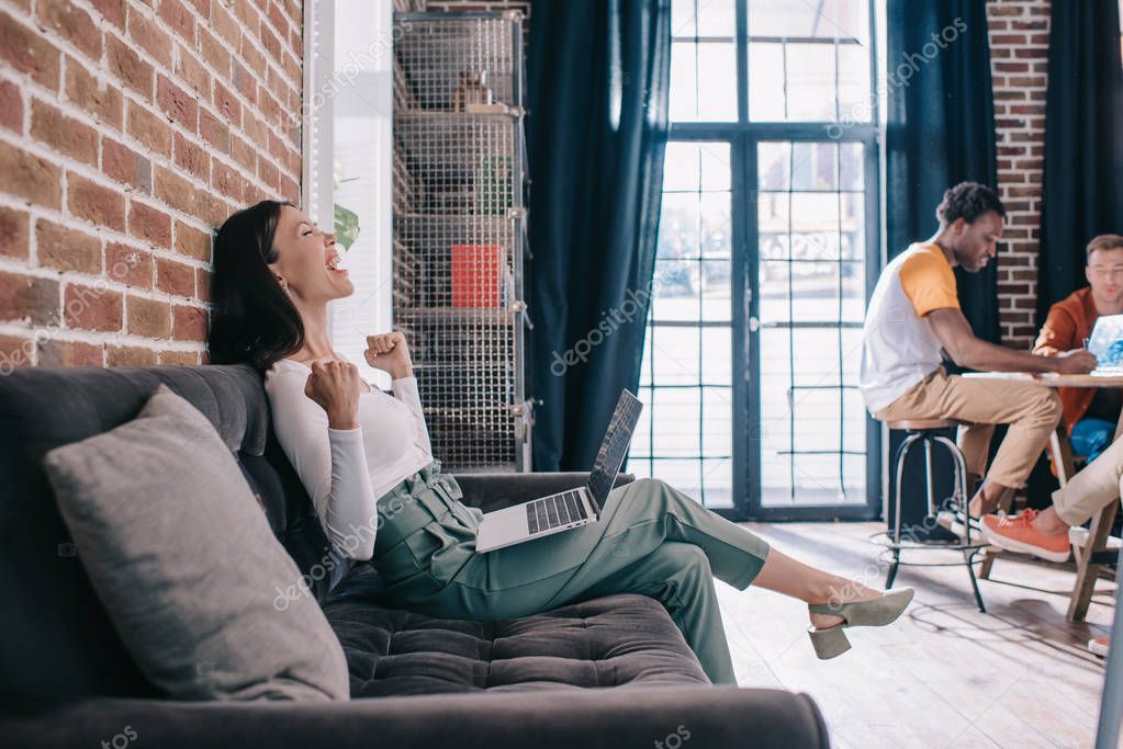excited businesswoman showing winner gesture while sitting on sofa near multicultural colleagues