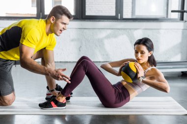 Sporcu kadın topla mekik çekiyor ve sporcu da spor merkezinde ona yardım ediyor.
