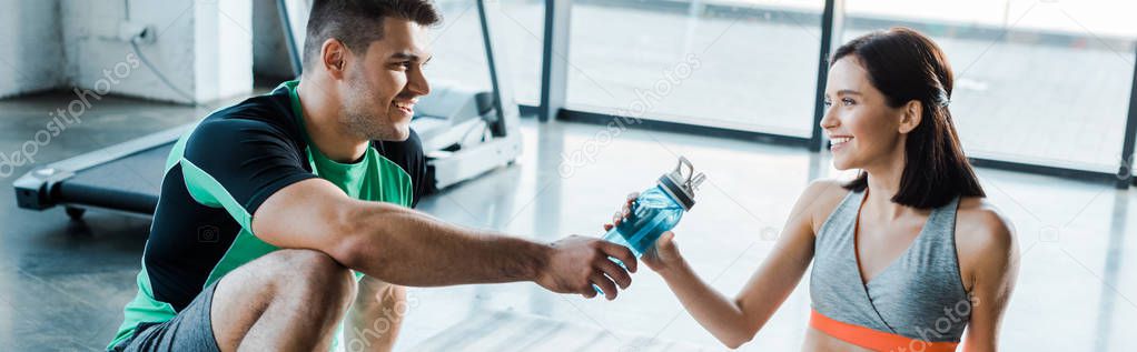 panoramic shot of smiling sportsman giving sports bottle to sportswoman in sports center