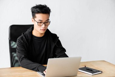 Gözlüklü Asyalı hacker masada oturuyor ve dizüstü bilgisayar kullanıyor. 