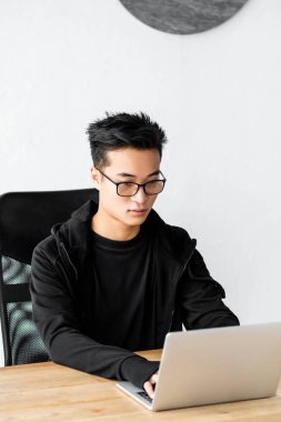 Gözlüklü Asyalı hacker masada oturuyor ve dizüstü bilgisayar kullanıyor. 