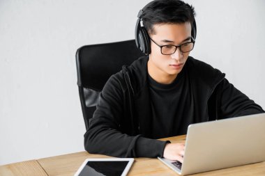 Kulaklıklı Asyalı hacker dizüstü bilgisayar kullanıyor ve masada oturuyor. 