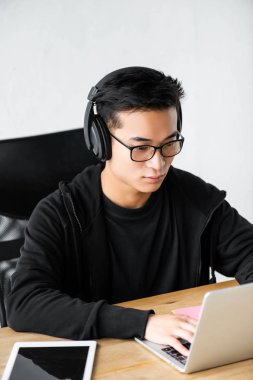 Kulaklıklı Asyalı hacker dizüstü bilgisayar kullanıyor ve masada oturuyor. 