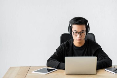 Kulaklıklı Asyalı hacker dizüstü bilgisayar kullanıyor ve masada oturuyor. 