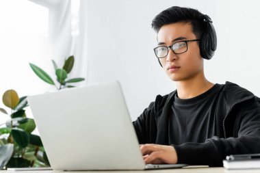 Kulaklıklı Asyalı hacker dizüstü bilgisayar kullanıyor ve masada oturuyor. 