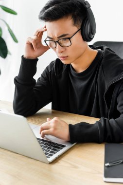 Düşünceli Asyalı hackerın yüksek açılı görüşü. Kulaklıklı, dizüstü bilgisayarlı ve masada oturuyor. 