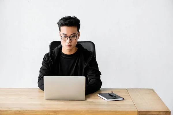 asian hacker in glasses sitting at table and using laptop