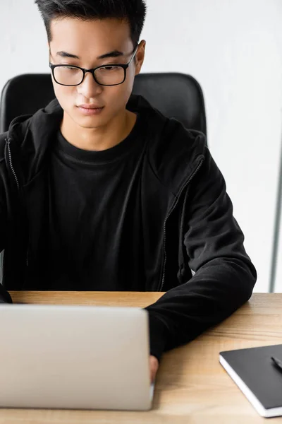 Azjatycki Haker Okularach Siedzi Przy Stole Pomocą Laptopa — Zdjęcie stockowe