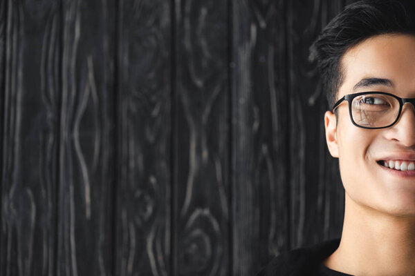 cropped view of smiling asian man in glasses looking away on wooden background 