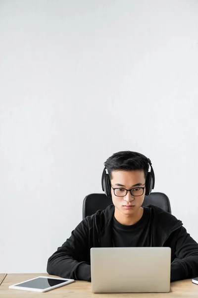 asian hacker in headphones using laptop and sitting at table
