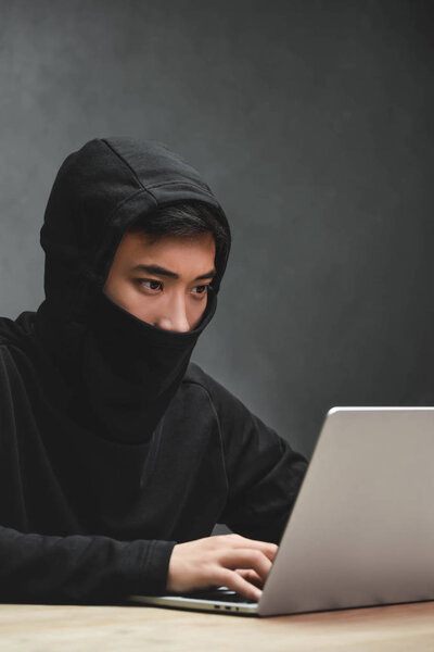 asian hacker with obscured face using laptop and sitting at table 
