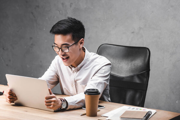 smiling asian seo manager sitting at table and holding laptop 