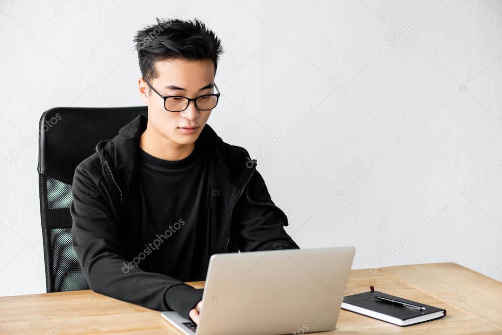 asian hacker in glasses sitting at table and using laptop 
