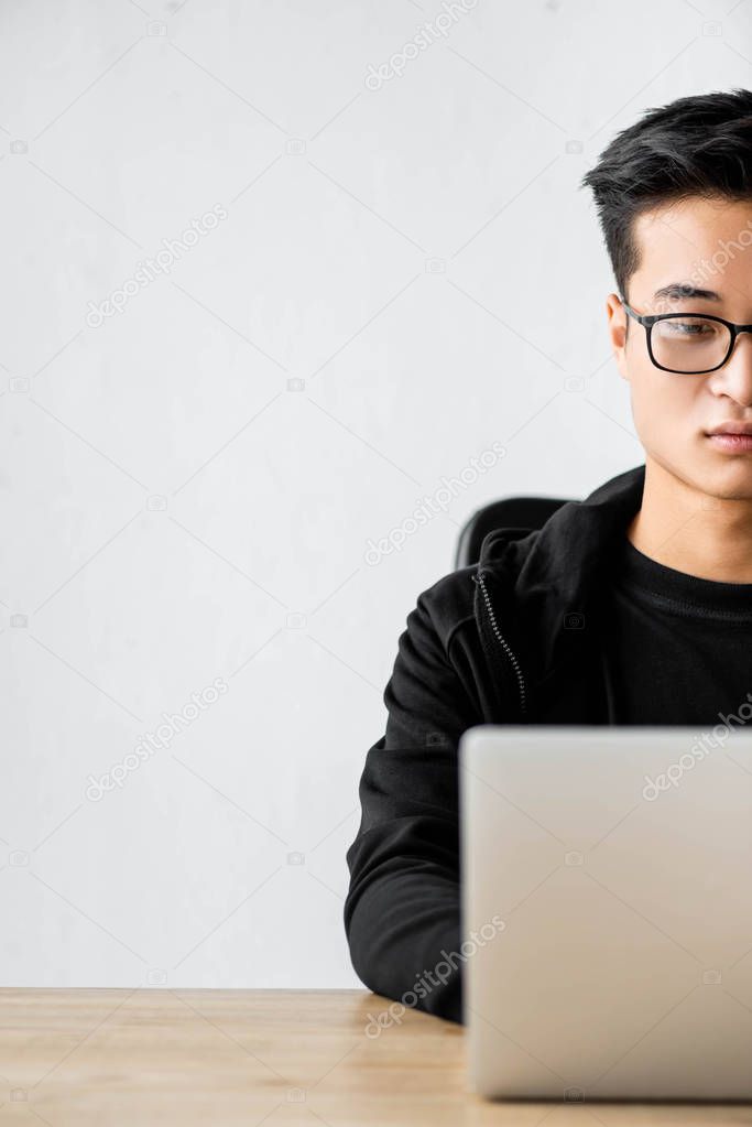 cropped view of asian hacker in glasses sitting at table and using laptop 