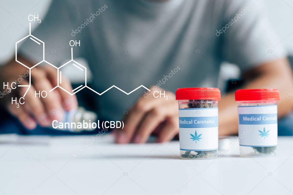 selective focus of bottles with medical cannabis on table with man on background with cbd molecule illustration