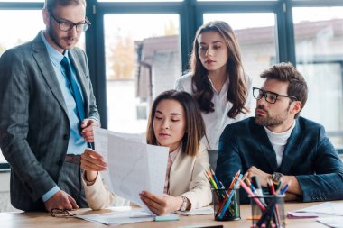 selective focus of businessmen looking at documents near multicultural businesswoman in office clipart