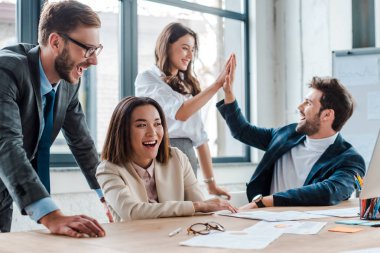 selective focus of happy businessman in glasses smiling near multicultural coworkers giving high five in office  clipart