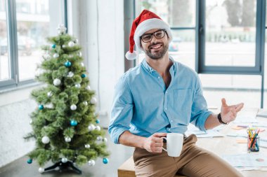 Noel Baba şapkalı ve bardaklı neşeli sakallı adamın seçici odak noktası. Ofisteki noel ağacının yanında kahve bardağıyla. 