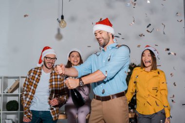 happy businessman in santa hat holding champagne bottle near coworkers and falling confetti  clipart